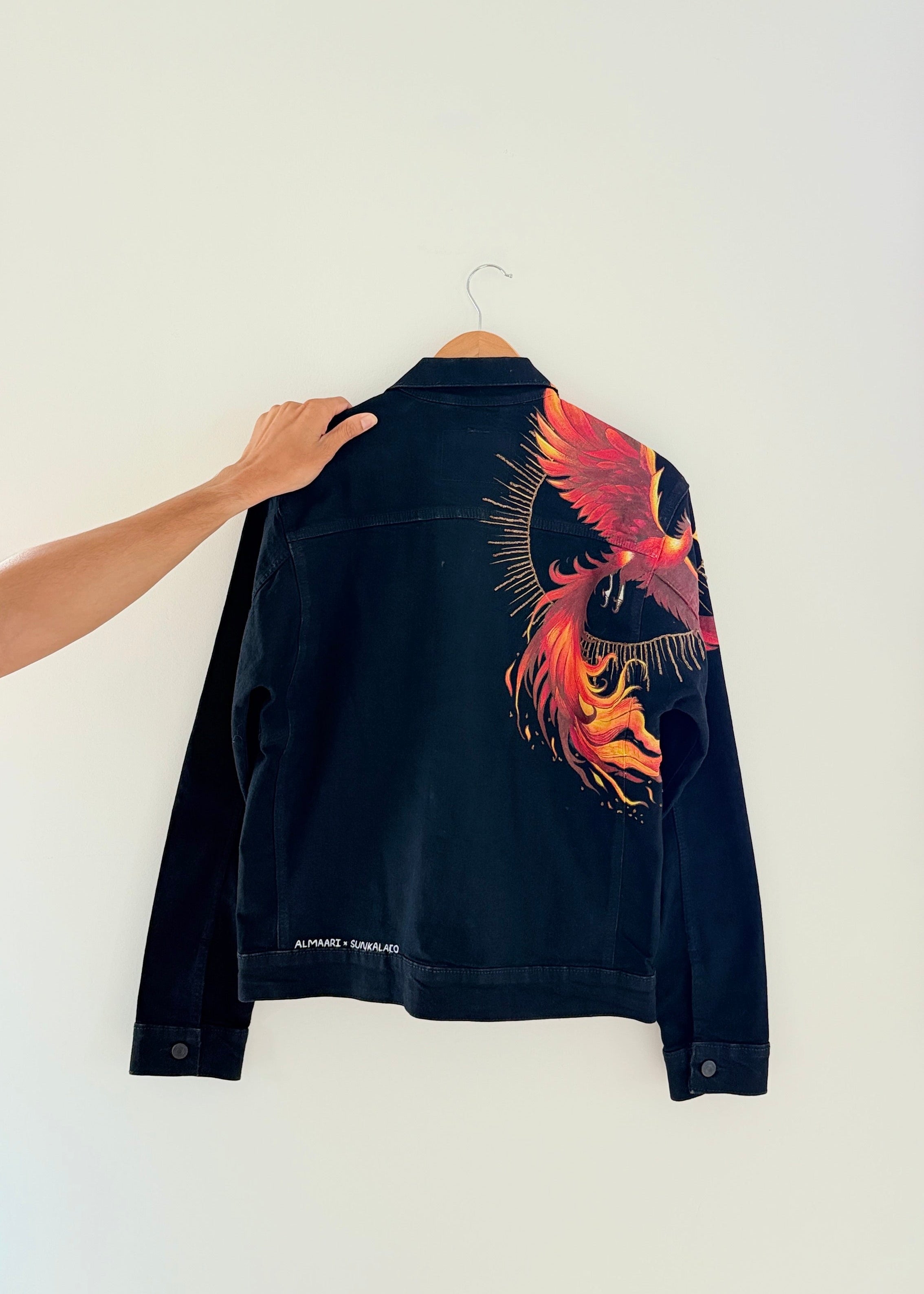 A black denim jacket with a large red, orange, and yellow phoenix graphic on the back, hanging on a wooden hanger against a plain white wall. A hand is holding the collar of the jacket.