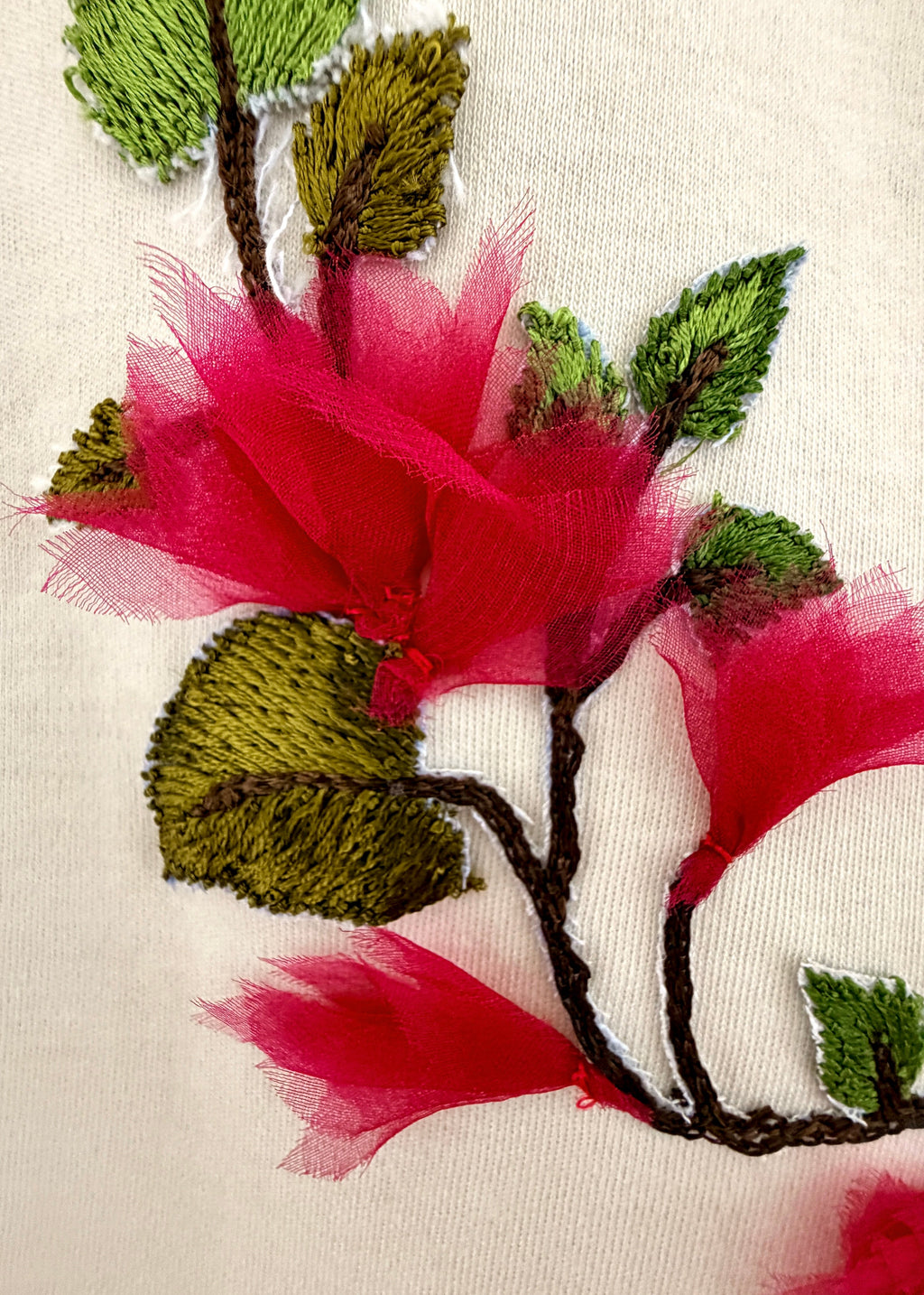 Close-up of embroidered flowers on a white fabric surface, featuring bright pink petals and green leaves with dark brown stems.