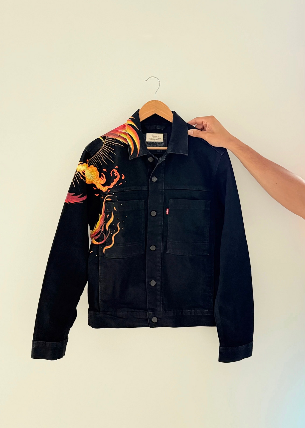 A black denim jacket with a vibrant orange, yellow, and red phoenix graphic printed on the right shoulder, hanging on a wooden hanger against a plain off-white wall. A hand is holding the collar of the jacket.